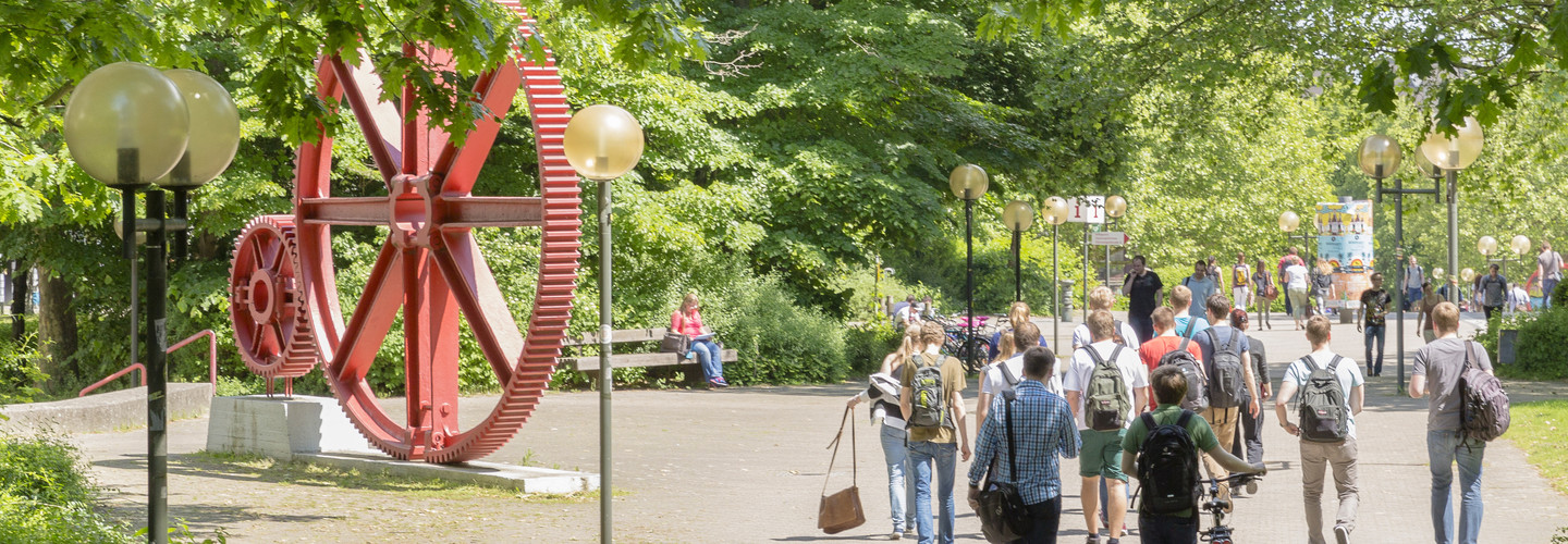 Studierende laufen über den Campus Nord. Es ist eine große rote Skulptur in Form von großen Zahnrädern zu erkennen.