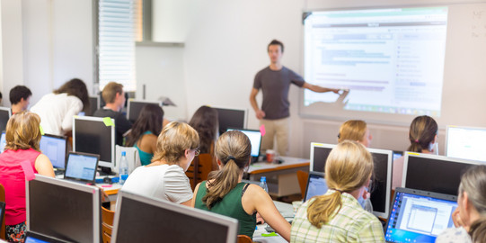 Computerunterricht Viele Menschen sitzen in einem Kursraum vor Computern. Vorne steht ein Lehrer und zeigt etwas auf einem Bildschirm.
