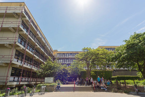 Chemiegebäude: Ein großes, breites, graues Gebäude mit Fensterfronten und Balkonen vor den Fenstern, die mit roten Metallstangen abgegrenzt werden. In der Mitte eine Treppe, auf der Menschen sitzen und links vor der Treppe Fahrräder in Fahrradständern.